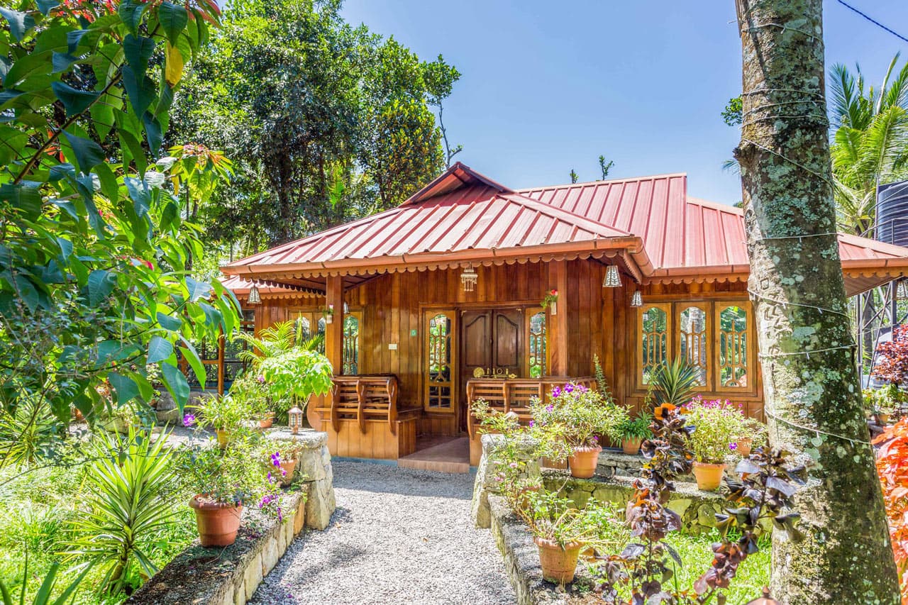 Wooden cottage with a red roof surrounded by lush tropical greenery and colorful potted flowers.